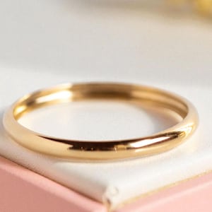 May include: A polished gold band ring. The ring has a smooth, rounded design and is likely made of gold. The ring is shown on a white and pink surface.