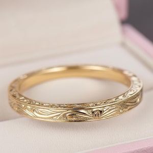 Può includere: Un anello d'oro con un intricato motivo floreale. L'anello è esposto all'interno di un portagioie rosa. Il design dell'anello presenta intagli dettagliati di foglie e altri elementi naturali, creando un'estetica delicata ed elegante.
