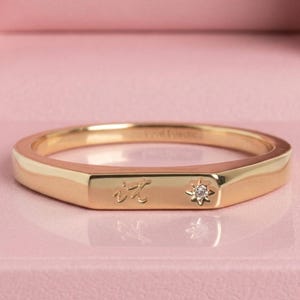 Può includere: Un anello d'oro dal design geometrico. L'anello presenta una piccola pietra a forma di diamante e un'iscrizione. L'anello è esposto in un portagioie rosa.