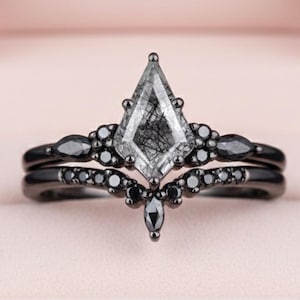 May include: A black metal ring set featuring a kite-shaped rutilated quartz centre stone. The band is accented with small black stones in a marquise and round cut, arranged in a symmetrical cluster design that follows the shape of the centre stone.