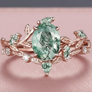 May include: A rose gold ring featuring an oval-cut green moss agate centre stone with dark inclusions. The band is designed with intricate metal leaf motifs and small, clear accent stones embedded along the branches surrounding the centre gem.
