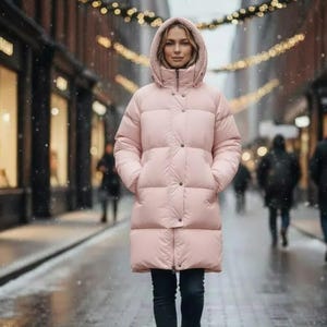 Pode incluir: Um casaco acolchoado rosa claro com capuz e fecho de botões. O casaco vai até ao joelho e tem bolsos. A pessoa está a usar calças pretas. O fundo é uma rua com neve com figuras desfocadas e montras.