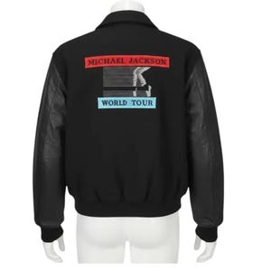 Könnte beinhalten: Schwarze Bomberjacke mit schwarzen Lederärmeln. Auf der Rückseite befindet sich ein rotes Rechteck mit "MICHAEL JACKSON" in weißem Text, eine Grafik eines Tänzers und ein blaues Rechteck mit "WORLD TOUR" in weißem Text.