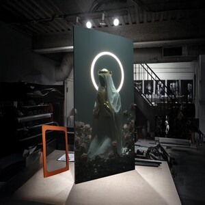 May include: A large, dark artwork featuring a figure with a glowing halo and gold accents, surrounded by roses. The figure is draped in white fabric, with hands clasped in prayer. An orange frame sits in front of the artwork.