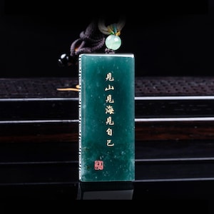 May include: A rectangular dark green jade pendant featuring vertical gold Chinese characters and a small red seal stamp at the bottom. The pendant has a white geometric border pattern along the side and is suspended from a cord with a light green bead.