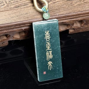 May include: A rectangular dark green jade pendant featuring vertical gold-colored Chinese characters and a small red square seal. The pendant is attached to a gold-toned metal loop, a small light green bead, and a thick dark brown braided cord.