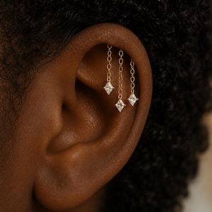 Könnte beinhalten: Nahaufnahme eines goldenen Ohrpiercings mit drei hängenden Ketten, an denen jeweils ein quadratischer, klarer Edelstein hängt. Der Schmuck ist vor dem Hintergrund eines Ohrs und dunklen Haares abgebildet.
