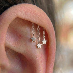 Puede incluir: Primer plano de una oreja con tres pendientes de cadena plateada, cada uno con un dije de estrella colgante. Los pendientes están sujetos al cartílago de la oreja, creando un efecto en capas.