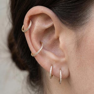 Op de afbeelding: Close-up van een oor versierd met meerdere gouden oorringen. De oorbellen zijn bezet met kleine, sprankelende steentjes. Eén oorbel is een helix-ring en twee zijn kleine ringen op de oorlel. De sieraden zijn tegen de huid geplaatst.