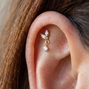 Puede incluir: Un piercing dorado con un diseño colgante. El piercing presenta tres piedras claras de talla marquesa dispuestas en forma de hoja, con una piedra redonda y clara colgando debajo. La joya está colocada sobre la piel de una oreja.
