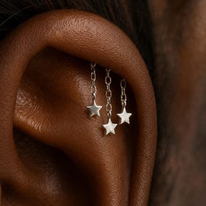 Può includere: Primo piano di un orecchino d'argento con tre ciondoli a forma di stella appesi a catenine. L'orecchino è posto contro un orecchio marrone. I ciondoli a forma di stella sono sospesi a delicate catenine, creando un effetto a strati.