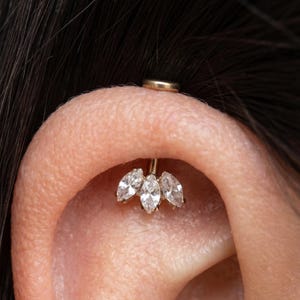 Op de afbeelding: Een goudkleurige oorbel met drie marquise-geslepen heldere edelstenen gerangschikt in een cluster. Een kleine, ronde gouden stud is zichtbaar aan de achterkant van het oor. De sieraden zijn geplaatst tegen de achtergrond van het oor en donker haar.