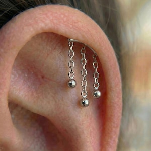 Può includere: Orecchino industriale a bilanciere argentato con tre catenelle pendenti, ciascuna terminante con una piccola sfera rotonda. L'orecchino è mostrato in un piercing all'orecchio, evidenziando il design.