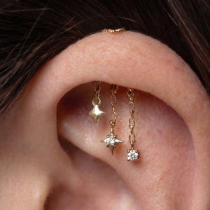 May include: Close-up of an ear with multiple gold earrings. One earring has a star charm, others dangle with chain links and gemstones. A small gold hoop is at the top of the ear.