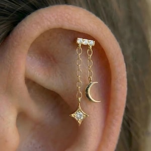 以下が含まれることがあります： ゴールドトーンのイヤリングピアス。小さなクリアストーンが付いたバーが、チェーンで三日月と中心にクリアストーンが付いた四芒星に繋がっています。イヤリングは耳に装着されています。