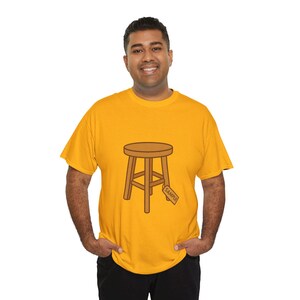Può includere: T-shirt giallo senape con un'illustrazione a cartoni animati di uno sgabello di legno. Lo sgabello ha una seduta rotonda e tre gambe. Una piccola etichetta con la scritta "SAMPLE" è attaccata allo sgabello. La maglietta è indossata da una persona.