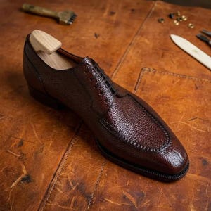 Peut inclure: Une chaussure richelieu en cuir marron foncé avec une texture grainée distincte, dotée d'un bout rapporté cousu, de lacets noirs fins et d'un embauchoir en bois clair inséré à l'intérieur pour maintenir la forme de la chaussure.