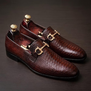 Pode incluir: Um par de mocassins castanho-escuros com um padrão texturizado de pele de cobra, um ornamento de metal dourado no peito do pé e formas de madeira escura com um puxador dourado inseridas para manter a forma.