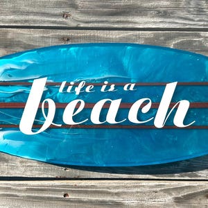 May include: A blue surfboard-shaped sign with the phrase "life is a beach" in white script. The sign has a blue and white wave design with three brown wooden stripes. The sign is displayed on a wooden background.