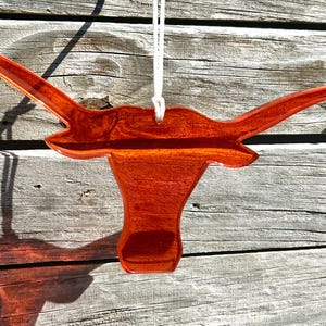 May include: A wooden ornament in the shape of a longhorn steer head, suspended by a white string. The ornament is a rich, reddish-brown color with a glossy finish. The background is weathered wood.