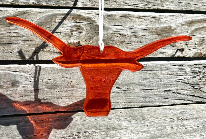 May include: A wooden ornament in the shape of a longhorn steer head, suspended by a white string. The ornament is a rich, reddish-brown color with a glossy finish. The background is weathered wood.