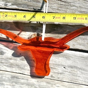 May include: A transparent orange longhorn ornament, approximately 10 inches wide, hangs from a white string. The ornament is made of a glossy material and is set against a weathered wood background. A yellow measuring tape is placed above the ornament.