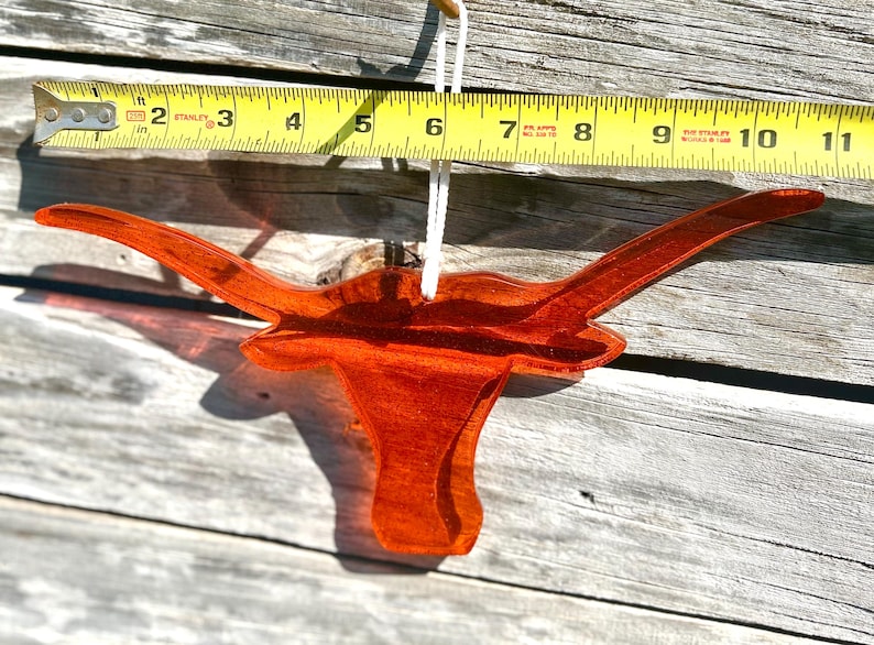 May include: A transparent orange longhorn ornament, approximately 10 inches wide, hangs from a white string. The ornament is made of a glossy material and is set against a weathered wood background. A yellow measuring tape is placed above the ornament.