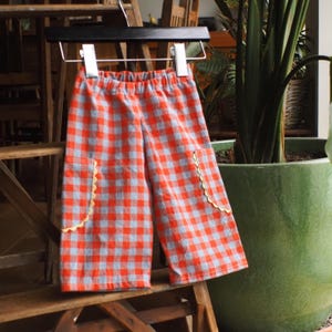 Könnte beinhalten: Eine Kinderhose mit rot-blauem Vichy-Karo-Muster. Die Hose hat zwei Vordertaschen mit gelben Zierkanten und einen elastischen Bund. Sie hängt an einem schwarzen Kleiderbügel.