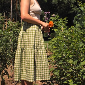 Op de afbeelding: Een midi-rok met een groen en wit gingham ruitpatroon. De rok heeft een gelaagd ontwerp met horizontale naden die een geplooid, vloeiend silhouet creëren. De stof oogt lichtgewicht met een zichtbare textuur over het gehele kledingstuk.
