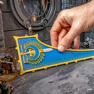 May include: A blue and gold decorative shield with a sword and laurel wreath design. The shield is held by a hand and is surrounded by miniature figurines, suggesting a tabletop game theme.