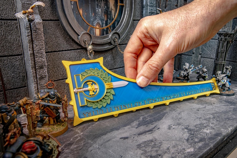 Peut inclure: Un bouclier d&eacute;coratif bleu et or avec une &eacute;p&eacute;e et une couronne de laurier. Le bouclier est tenu par une main et est entour&eacute; de figurines miniatures, sugg&eacute;rant un th&egrave;me de jeu de soci&eacute;t&eacute;.