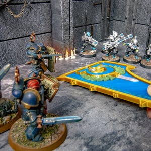 May include: Miniature figurines of armoured soldiers with swords and shields are arranged on a grey surface. A blue and gold decorative piece is held by a hand. The scene is set in a grey, textured environment.