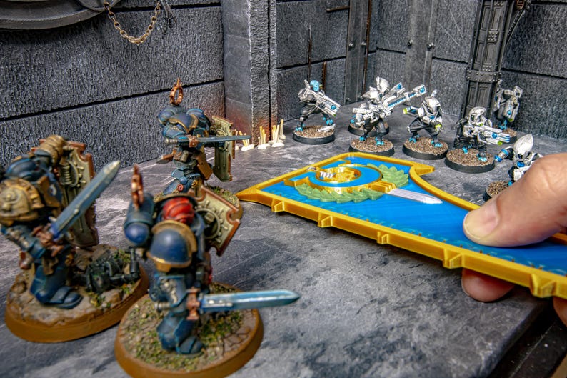 Peut inclure: Figurines miniatures de soldats blind&eacute;s avec &eacute;p&eacute;es et boucliers dispos&eacute;es sur une surface grise. Une pi&egrave;ce d&eacute;corative bleue et dor&eacute;e est tenue par une main. La sc&egrave;ne se d&eacute;roule dans un environnement gris textur&eacute;.