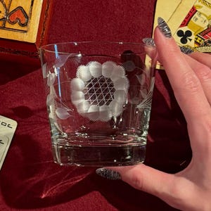 Könnte beinhalten: Ein klares, geätztes Glas mit floralem Design. Das Glas hat eine zentrale Blume mit einem geometrischen Muster und zarten Blattakzenten. Spielkarten und eine Holzkiste befinden sich im Hintergrund und deuten auf ein Spiel- oder Unterhaltungsthema hin.