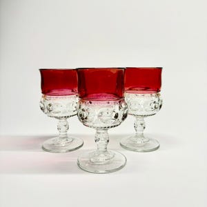 May include: Three vintage wine glasses with ruby red bowls and clear, patterned stems. The glasses are arranged on a white surface, showcasing their elegant design. The stemware is ideal for serving wine or other beverages.