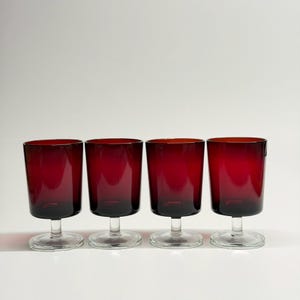 May include: Four ruby red wine glasses with a short stem and a rounded bowl. The glasses are made of a transparent material. The glasses are arranged in a row against a white background. Each glass is approximately 10 cm tall.