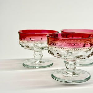 May include: Set of three clear glass dessert bowls with ruby red rims and decorative detailing. The bowls have a clear, textured base and stem. The red color fades into a lighter pink towards the center of the bowl.