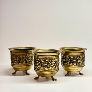 May include: Three antique brass planters with floral relief patterns. Each planter has a round shape, a wide rim, and three small, claw-shaped feet. The planters are a warm gold colour, with dark accents in the floral details.