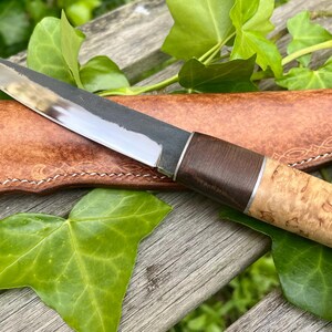 May include: A handcrafted knife with a polished steel blade and a wooden handle with a light brown and dark brown color scheme. The knife rests on a brown leather sheath with white stitching, surrounded by green leaves.