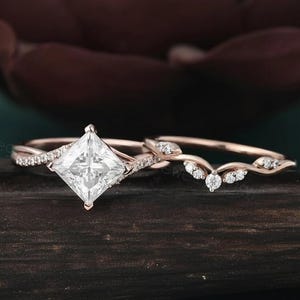 Op de afbeelding: Rosegouden verlovingsring set. De verlovingsring heeft een princess-cut middensteen en een delicate band. De trouwring heeft een gebogen ontwerp met kleine accentstenen. De ringen worden gepresenteerd op een donker houten oppervlak.