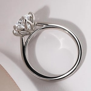 May include: A silver ring with a large, clear gemstone. The gemstone is held in place by multiple prongs, and the band is smooth and polished. The ring is set against a white background, with a shadow to the side.