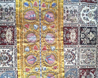 May include: A rectangular, golden-yellow textile with an embroidered floral design. The central motif features symmetrical pink and blue flowers, with a blue and white central stem. The border is a lighter gold, and the background is a patterned rug.