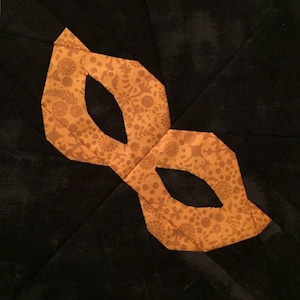 May include: A black fabric background with a gold patterned masquerade mask. The mask has two eye holes and is shaped like a teardrop.