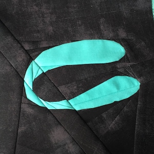 May include: A close-up of a teal blue fabric design on a black background. The fabric design is shaped like a curved letter C.
