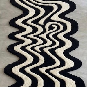 May include: A long, narrow runner rug featuring a high-contrast black and off-white pattern of fluid, wavy, and swirling lines. The rug has an irregular, organic shape with curved edges that follow the undulating design of the tufted surface.