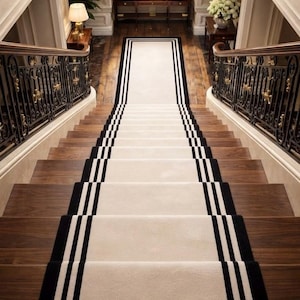May include: A long, rectangular stair runner carpet featuring a cream-colored center with bold black border stripes. The runner is installed down the center of a wooden staircase, flanked by dark metal railings with gold accents.