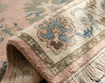 May include: A woven area rug featuring a soft pink base with intricate floral patterns in shades of blue, green, and cream. The rug has a thick, textured pile, a decorative border with repeating motifs, and long cream-colored fringe along the edge.