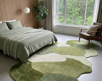 May include: An area rug featuring an irregular, organic shape with a multi-tonal pattern of sage green, olive green, and cream sections. The rug has a plush, textured pile and is positioned on a light gray tiled floor in a bedroom setting.