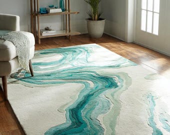 Puede incluir: Una alfombra grande con un diseño abstracto y fluido en tonos turquesa y verde sobre un fondo crema. El patrón evoca un río o un elemento acuático, añadiendo un toque natural a la decoración de la habitación.