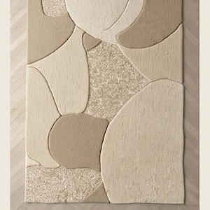 May include: Beige area rug with an abstract design. The rug features a pattern of overlapping shapes in varying shades of beige and tan. The rug has a textured surface and the word "Beige" is printed at the bottom.